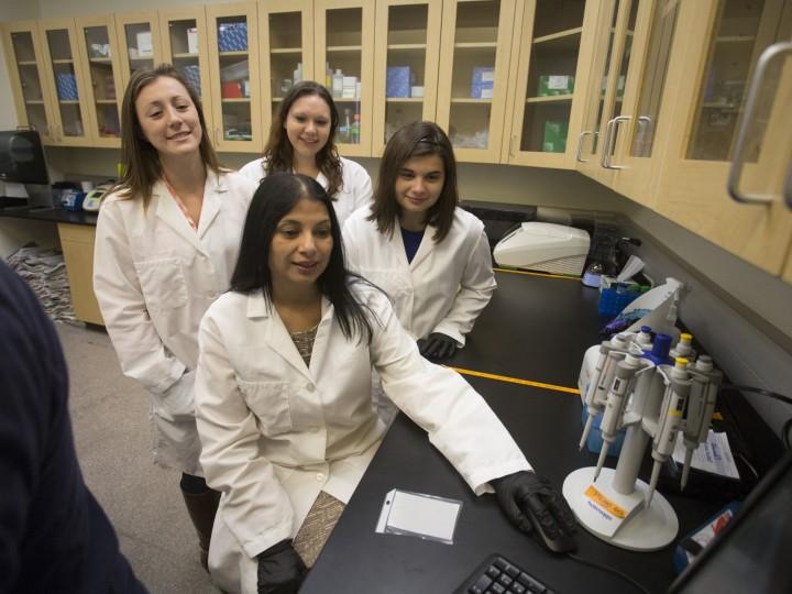 smiling students standing behind Dr.Malik in the lab while using a pipette